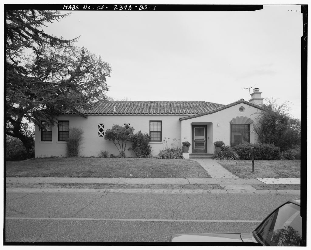 A single-storey house has a tiled roof, recessed entrance and small patterned openings, with trees at the left and a car corner in front.