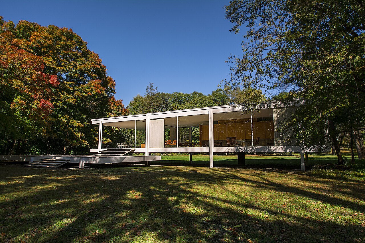 Farnsworth House seen through trees as a raised white steel frame enclosed by large clear glass panes.