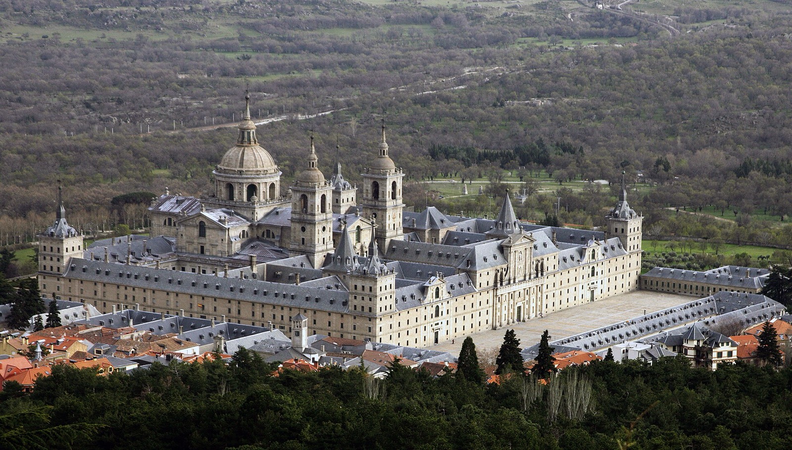 Oblique El Escorial view with long outer stone walls, western forecourt, church dome, roofs and surrounding settlement.
