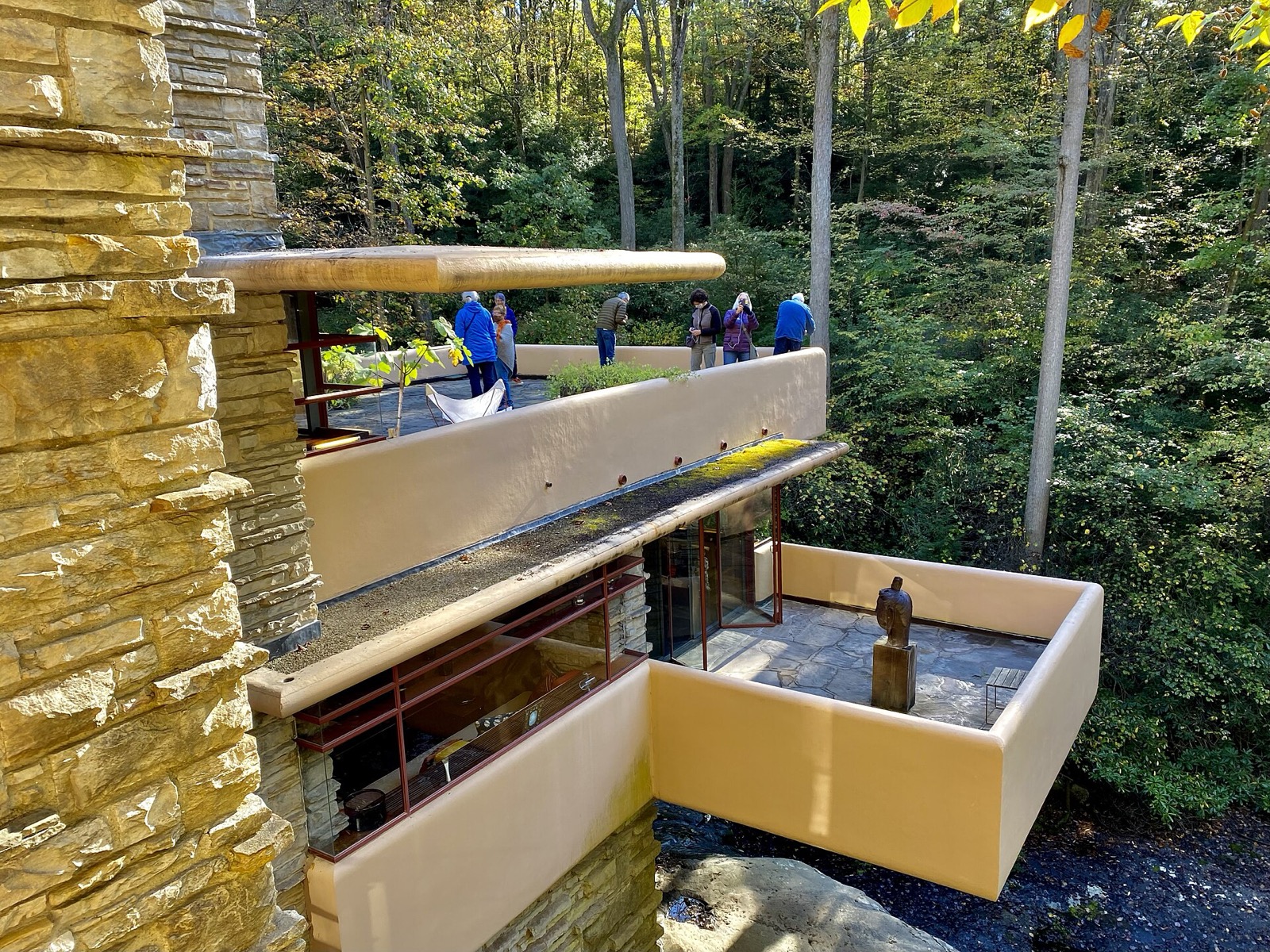 Terrace-level Fallingwater view with stacked projecting concrete roof edges beside sandstone chimney core and wooded slope.