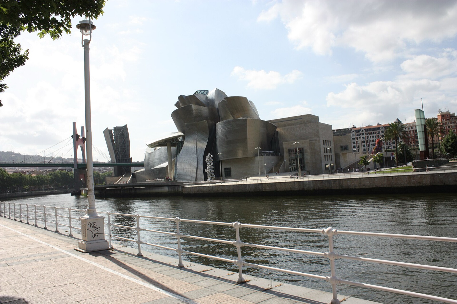 Guggenheim Bilbao across the Nervión River with curved titanium-clad masses, adjacent limestone block and bridge.