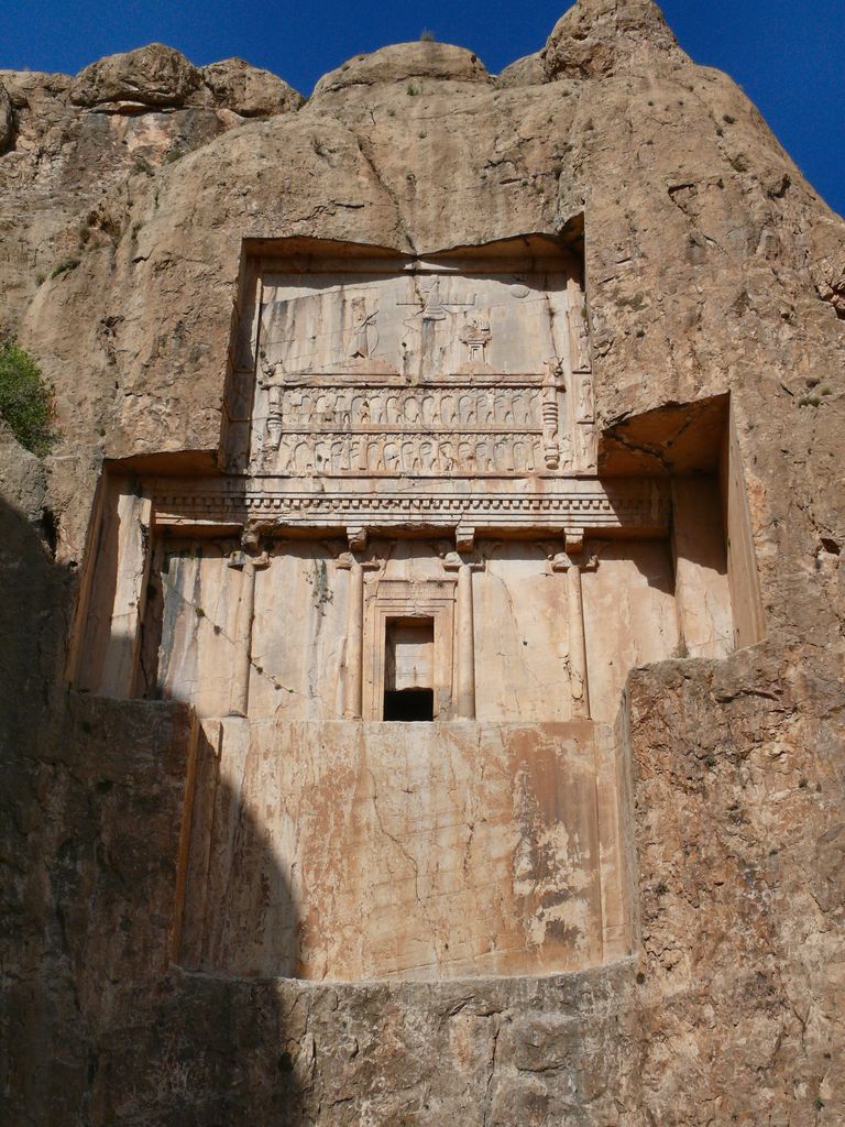 A cross-shaped carved tomb façade with engaged columns, figures and a dark central doorway is cut high into a rough cliff.
