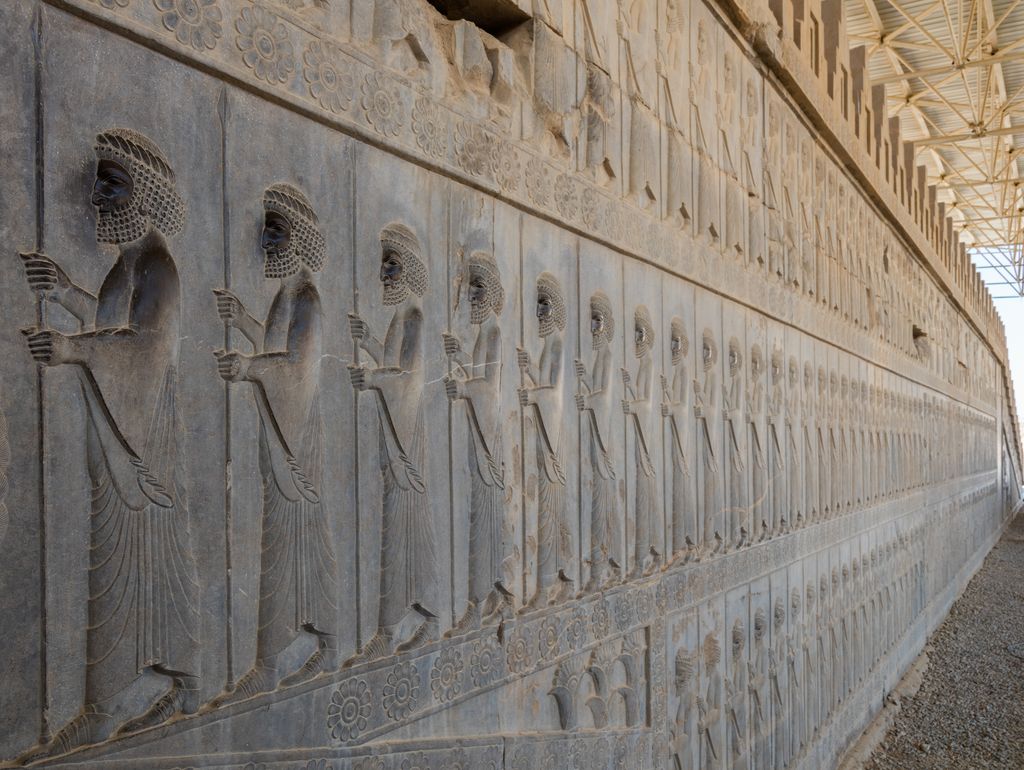 A long row of shallow stone relief figures holding spears recedes along a protected stair wall.