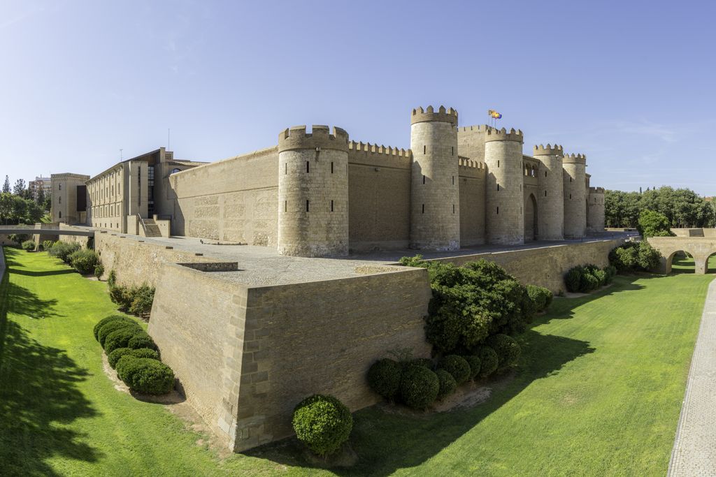 A pale rectangular fortified palace with rounded towers stands behind a ditch and green lawns in an urban setting.