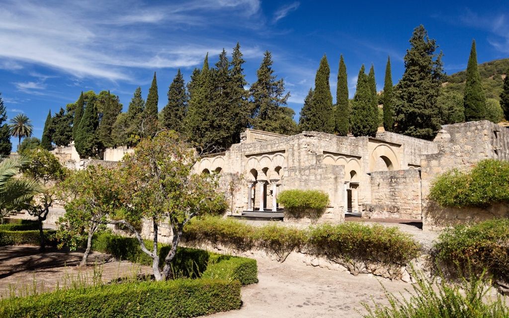 Low excavated stone walls and planted terraces lead to a partly reconstructed three-arched façade beneath cypress trees.