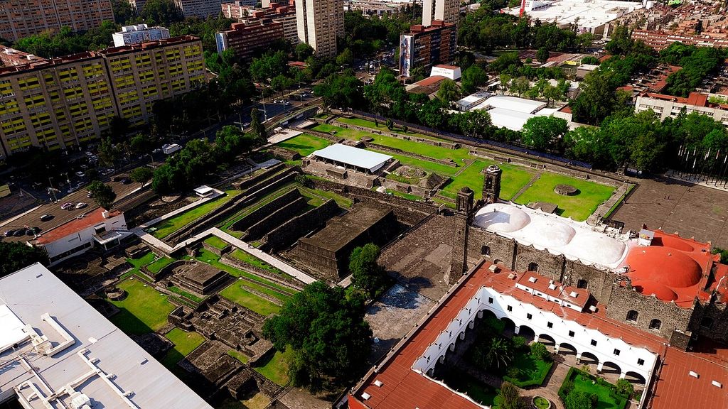 An aerial view shows excavated stone platforms beside a large historic church and cloister, surrounded by roads, trees and modern apartment towers.