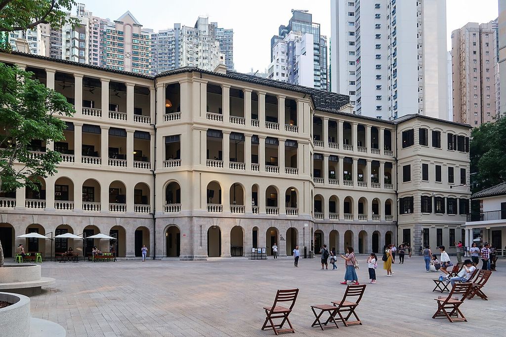 Four levels of arcades and open verandas face a paved courtyard with visitors and seats amid taller modern buildings.