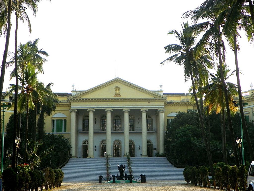 Tall columns and a pediment rise above broad entrance steps between palms and curving side ranges.