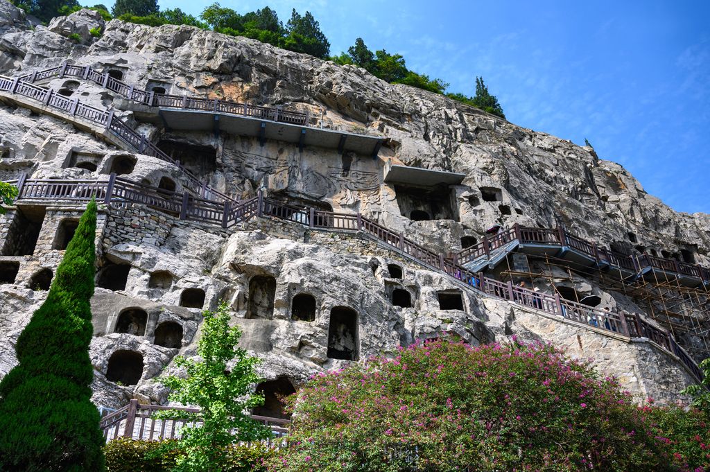A pale limestone cliff densely cut with caves and small niches rises behind zigzagging visitor stairs and railings.