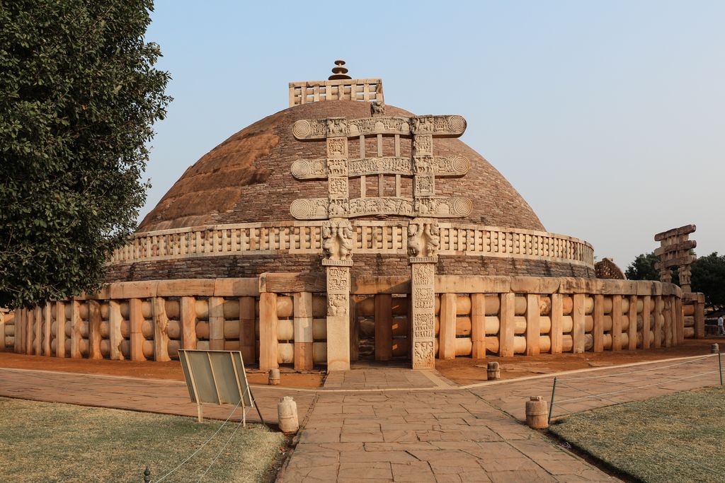 A large rounded stone-faced stupa stands behind a carved gateway and railing, with trees at either side.