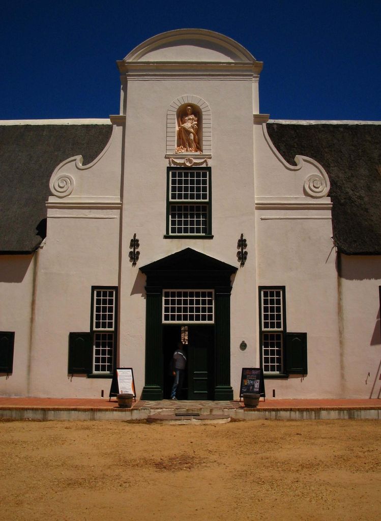 A curved pale gable with a sculpture niche and central window rises above the entrance and thatched roof.