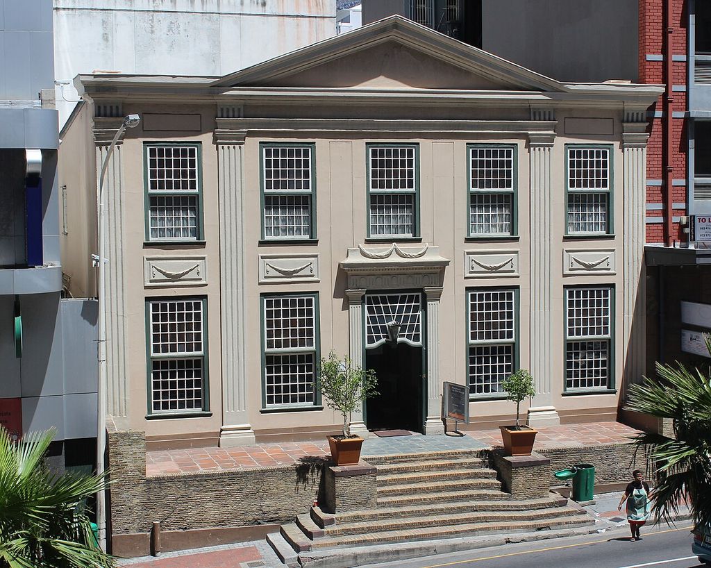A symmetrical two-storey frontage with pilasters and a pediment stands above a raised stoep and entrance steps.