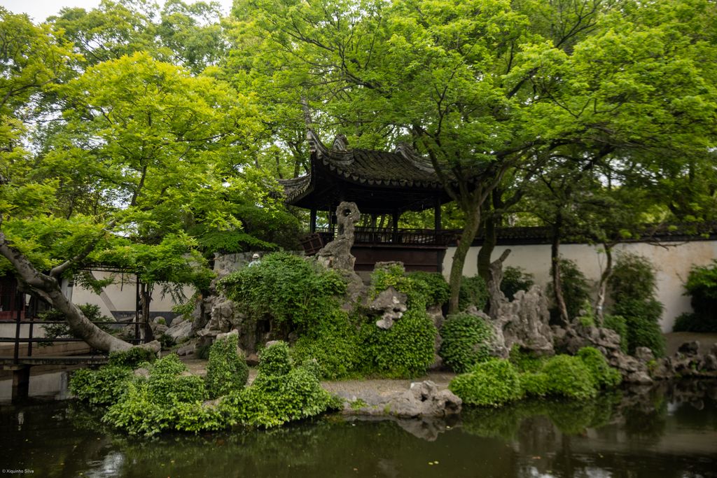 A shaded garden pond reflects dense trees, pale rockery, a small pavilion and covered walkway.