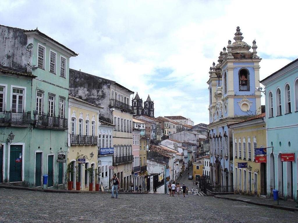 A sloping cobbled square descends between narrow multistorey houses and a blue church with twin towers.