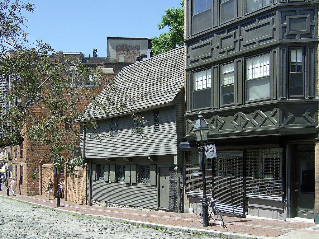 A low clapboard house with an overhanging upper floor and pitched roof stands between taller brick and decorated façades.