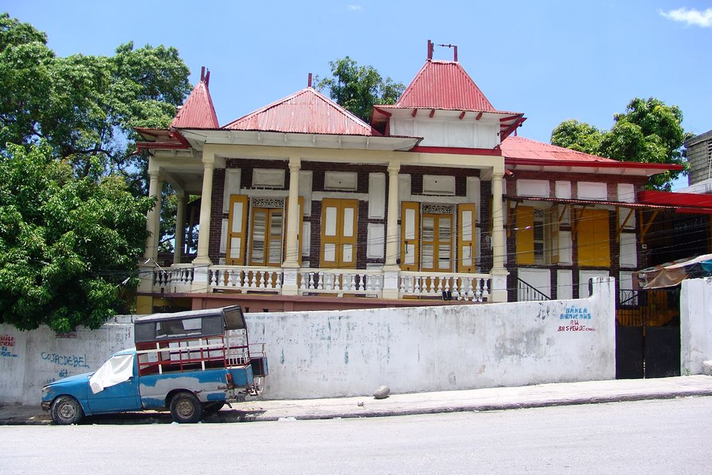 A raised one-storey house has a deep columned veranda, tall yellow shutters, timber fretwork and several steep red metal roofs behind a street wall.