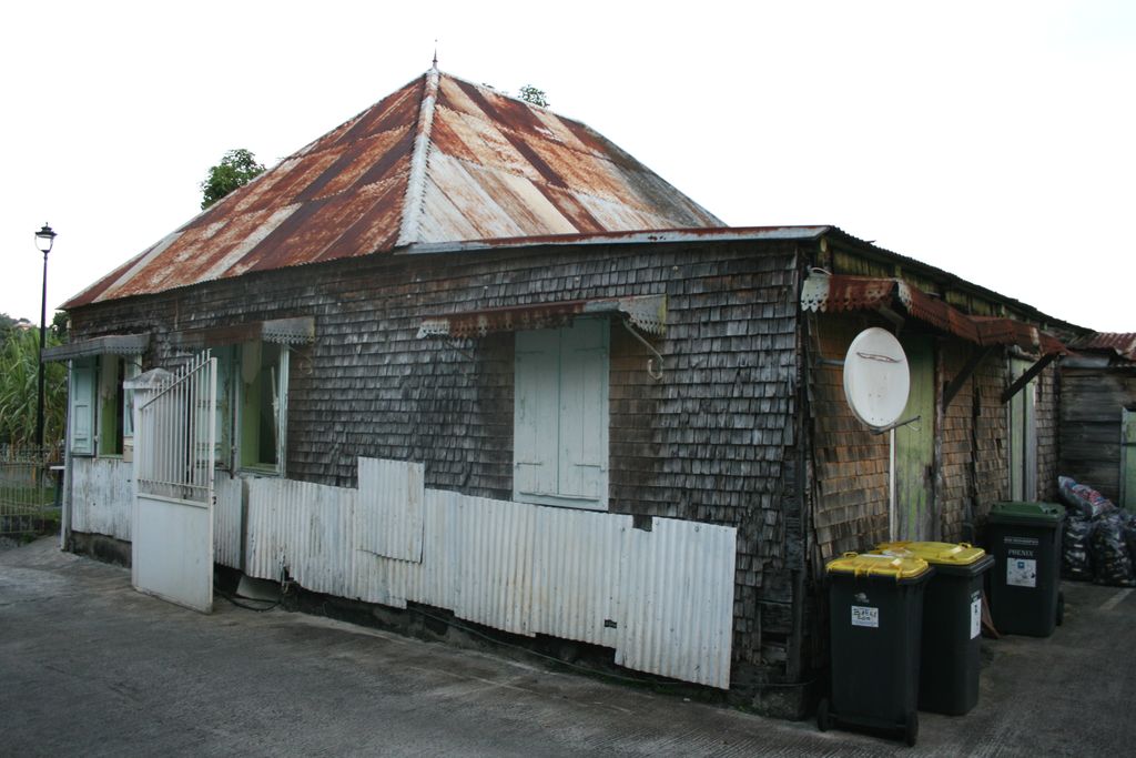 An oblique view shows a low timber-shingled house with a broad rusted hipped metal roof, narrow veranda, decorative roof-edge trim and modern bins and satellite dish.
