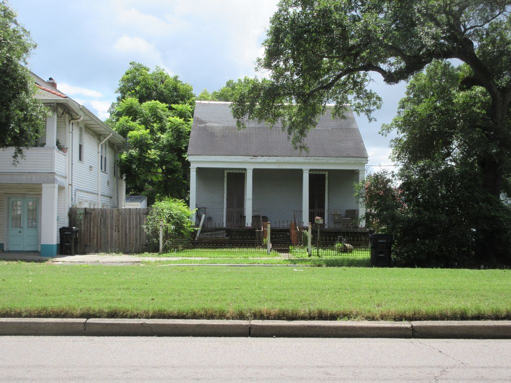 A low grey timber cottage with a steep roof has four openings across its street front and stands close to the pavement beneath large trees.