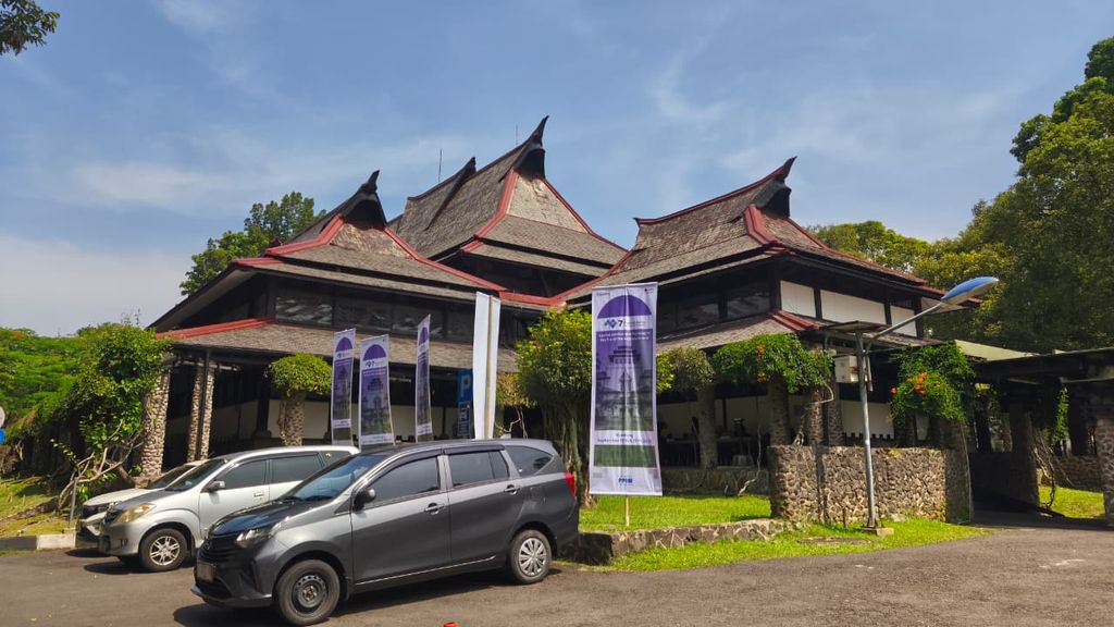 A large layered roof with sweeping ends rises above an institutional entrance, with cars and signs in the foreground.