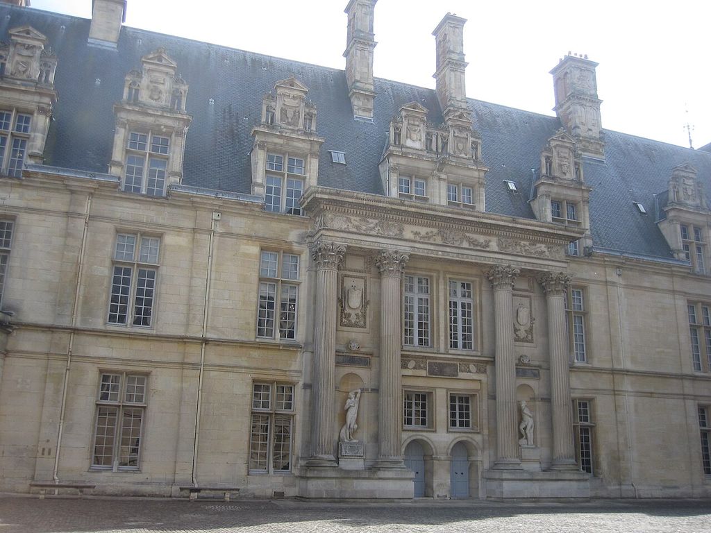 A long pale stone courtyard façade has a central columned portal with sculpture, rows of windows, ornate dormers, tall chimneys and a steep dark slate roof.