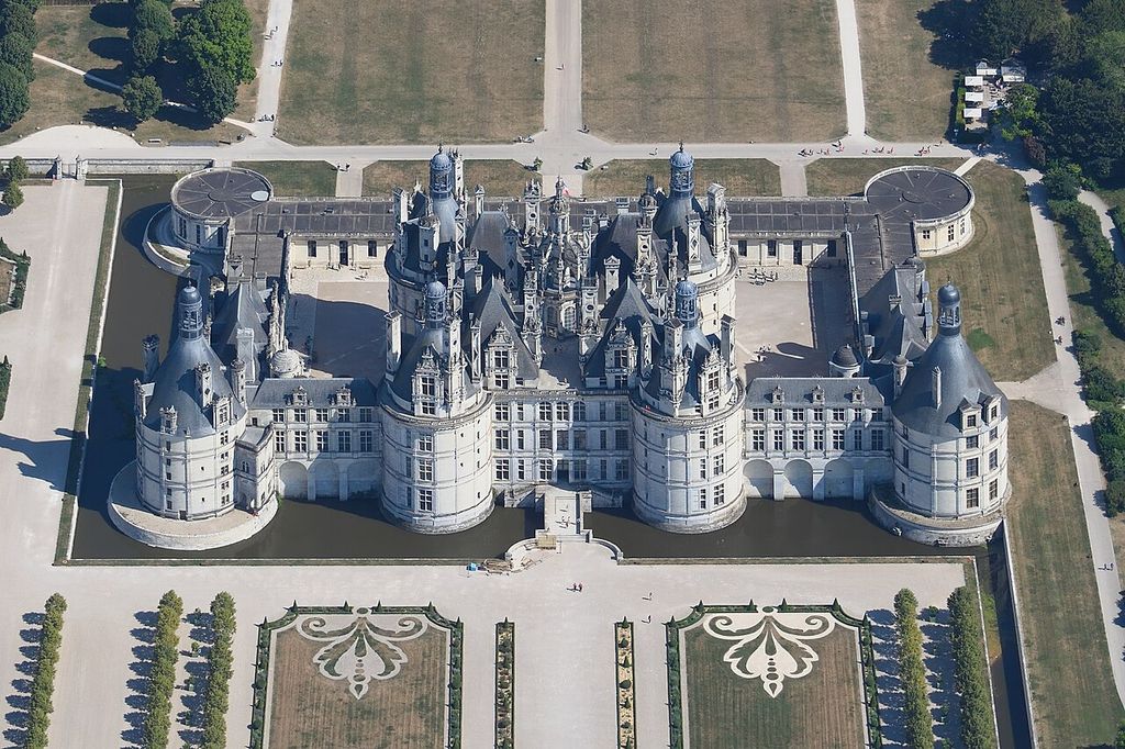 A near-vertical aerial view shows a vast pale château with a square central block, four round corner towers, crowded roofs, courts, moat and formal gardens.