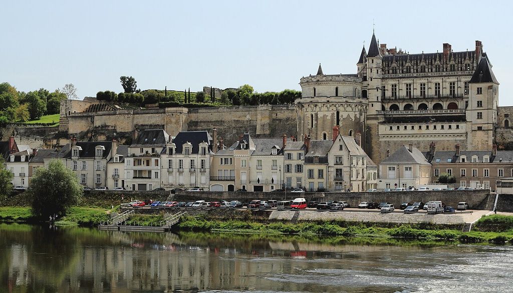 A pale château with a large round tower stands on a high stone terrace above riverside town houses, parked cars and the Loire.