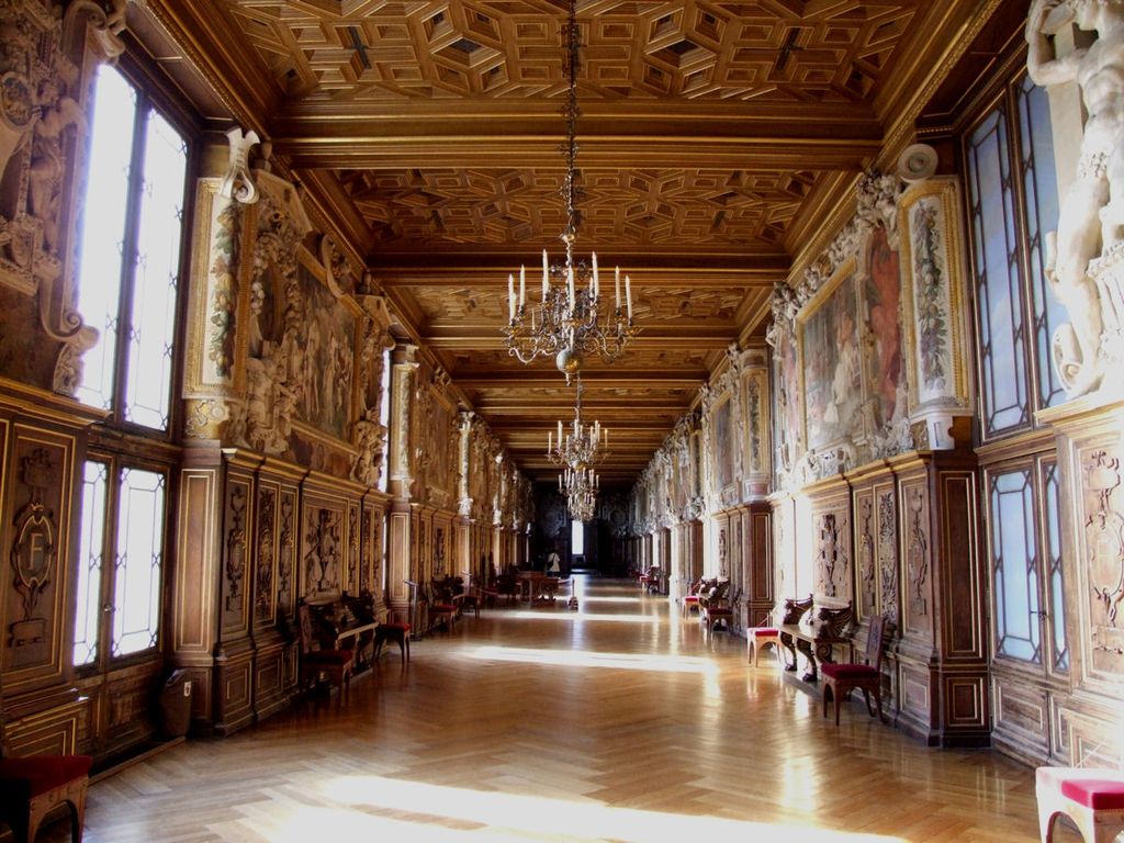 A long palace gallery has an elaborate coffered wood ceiling, polished floor, tall windows, chandeliers and walls combining dark panelling, paintings and pale sculpted frames.