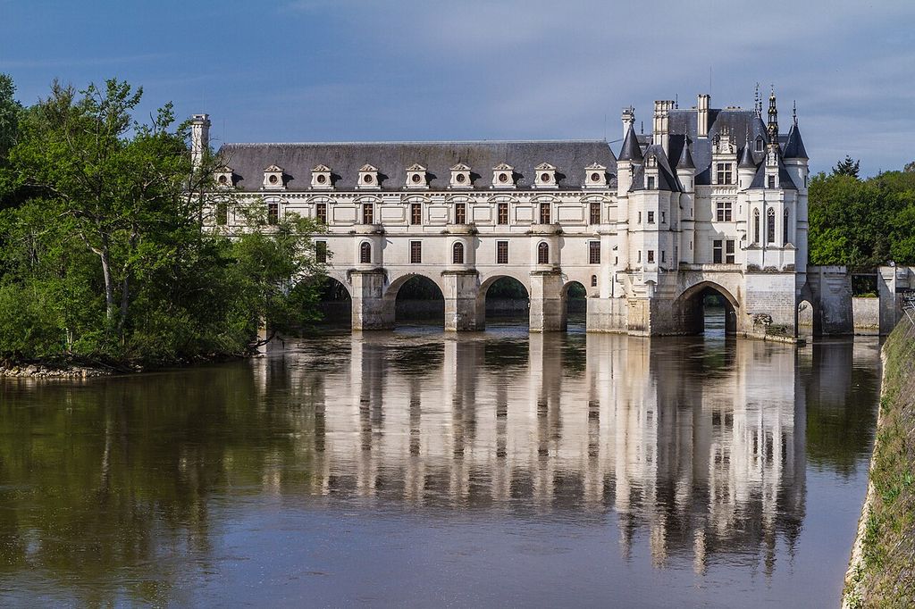 A pale château with turrets stands beside a long two-storey gallery carried across a broad river on five large stone arches.