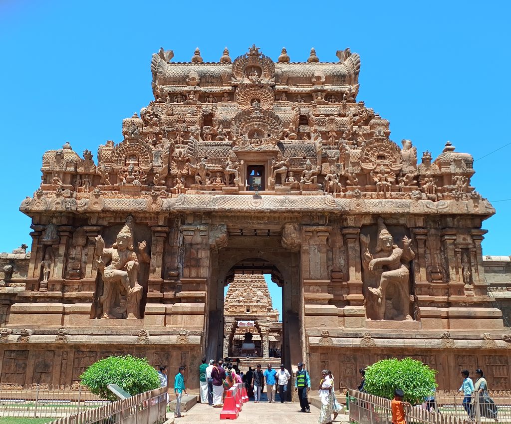 A richly carved stone gateway with a tall tiered crown and large guardians frames a passage into a temple complex.