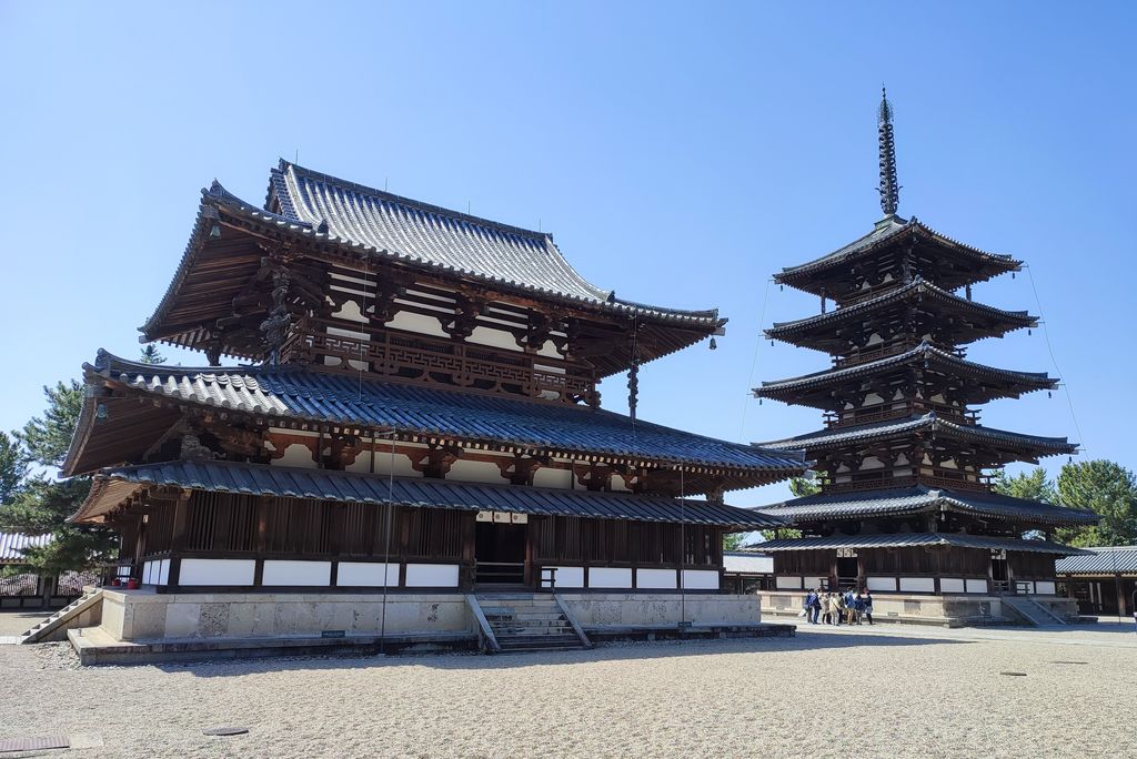 A dark timber worship hall and tall five-storey pagoda stand side by side across a pale gravel court beneath a blue sky.