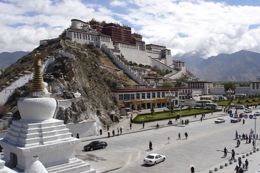 A vast white and red palace complex steps up a rocky mountain above a broad city square, roads and distant peaks.