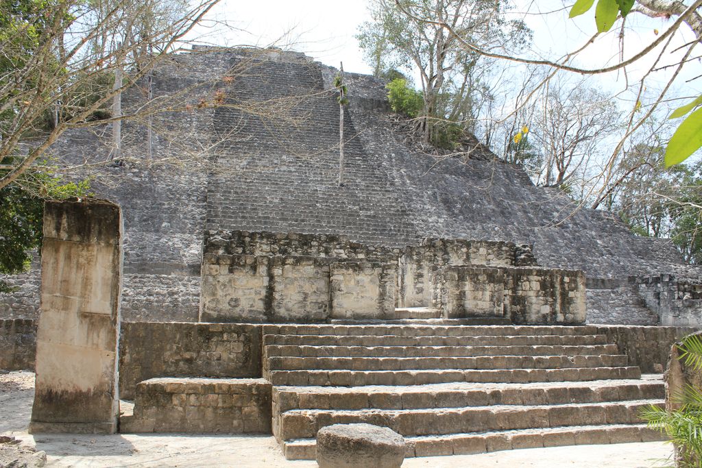 A broad weathered stone pyramid with a central stair rises among dense green trees under a pale blue sky.