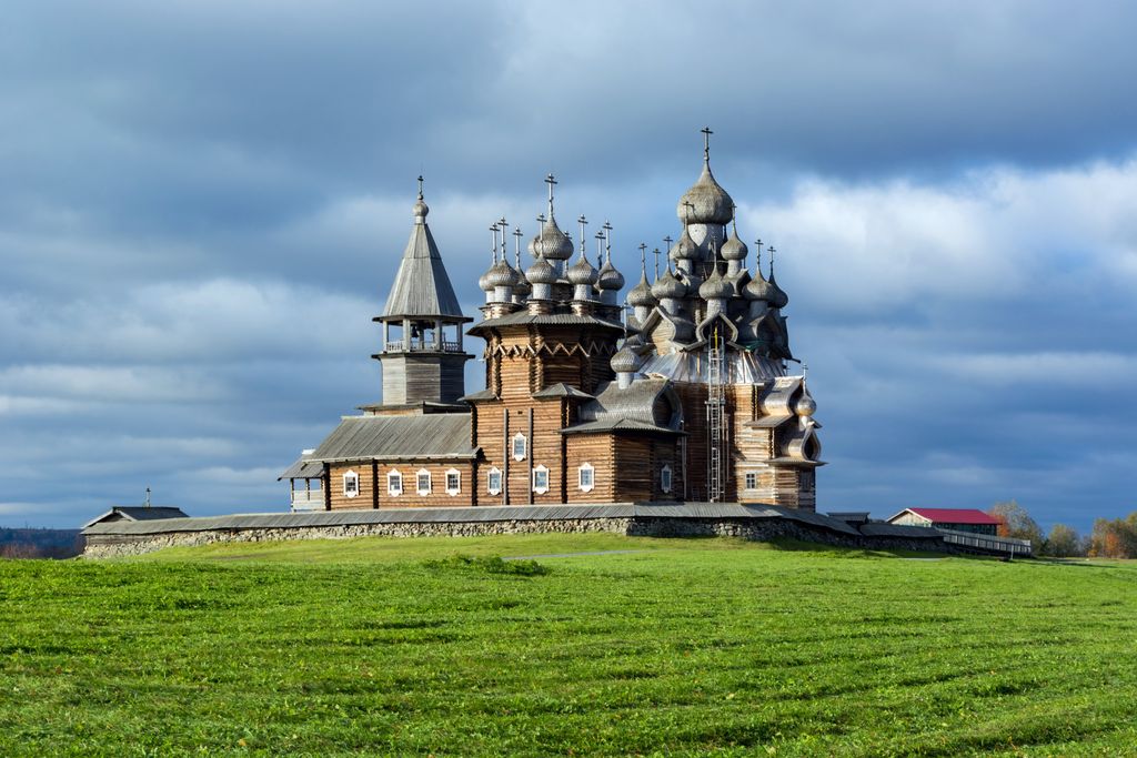 A tall wooden church crowned by many silver-grey onion domes stands in a green field beneath dark clouds.