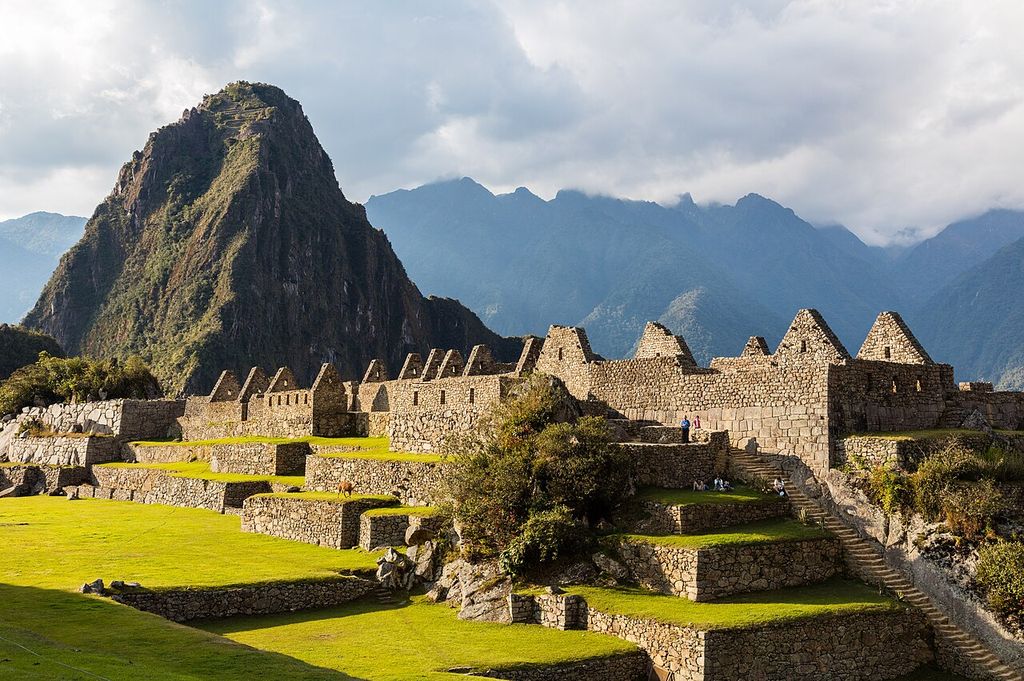Sunlit roofless stone houses and grass terraces step across a mountain ridge, with steep Huayna Picchu and clouded peaks beyond.