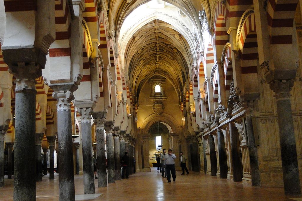 Red-and-white arches and dark columns meet a later high vaulted aisle with carved stone walls and visitors.