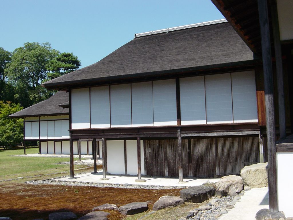 A raised timber building with pale sliding panels, dark boarded base and deep roof stands beside a carefully maintained garden.