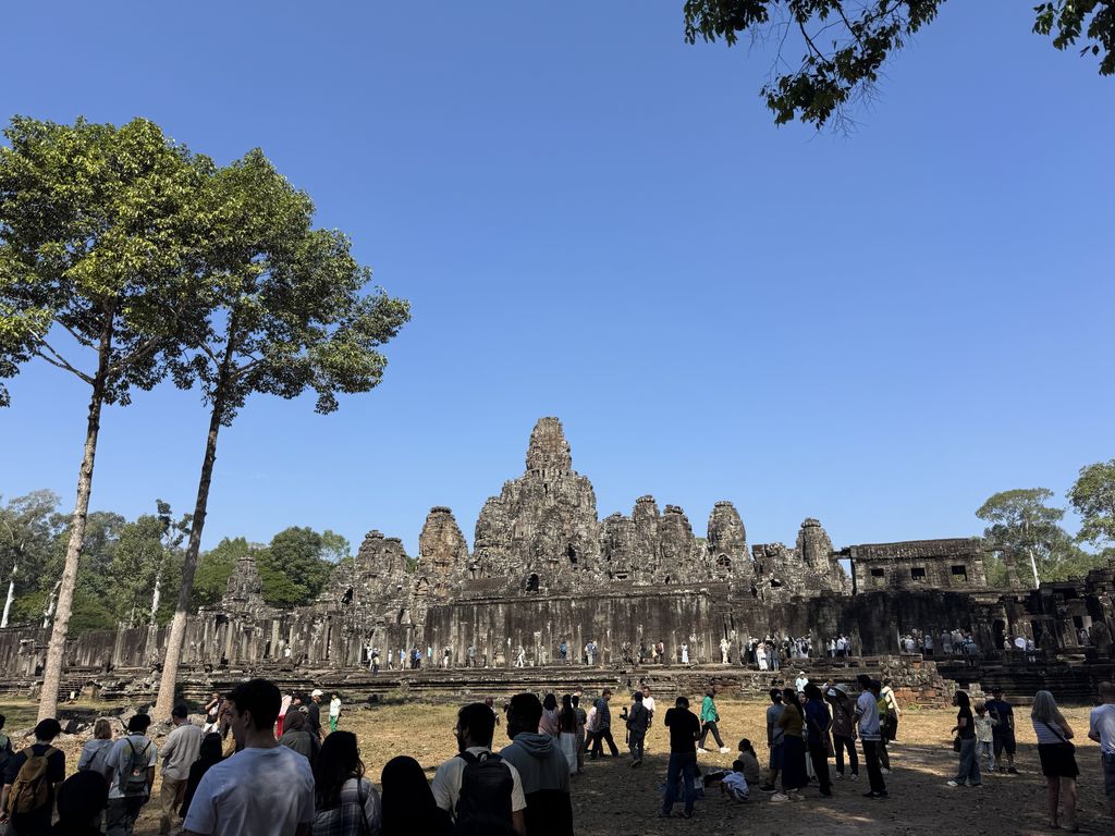A dense cluster of weathered stone towers with large carved faces rises above a gallery and a crowd of visitors.