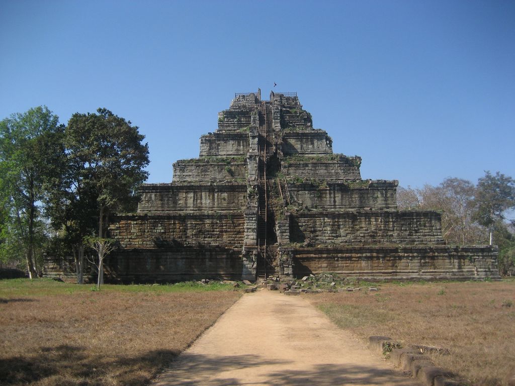 A seven-tiered weathered stone pyramid rises at the end of a sandy path with trees and lower ruins around it.