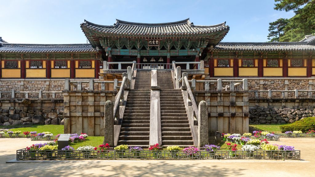 Twin stone stairways rise through terraces to a richly painted timber gate beneath grey tiled roofs and blue sky.