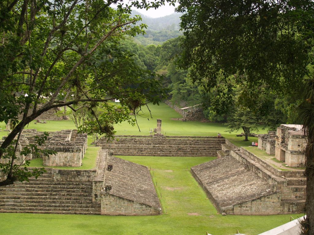 A long grass court runs between two sloping stone playing walls toward stepped masonry, framed by dense trees.