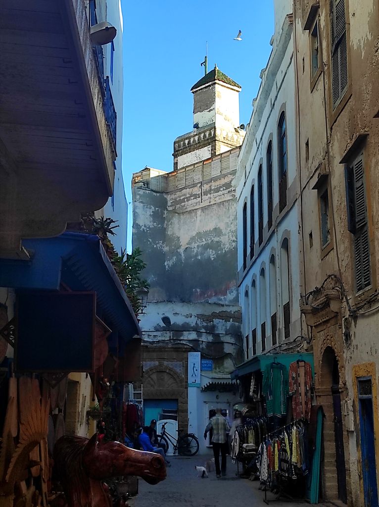 A narrow market street lined by worn white and ochre buildings leads to a tall square fortified tower under blue sky.