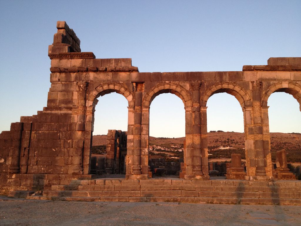 A warm stone arcade with four large openings stands among low excavated ruins beneath a pale blue sky.