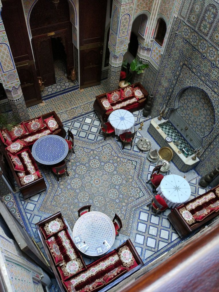 A high view into a tiled courtyard with round tables, red banquettes, carved plaster and dark timber galleries.