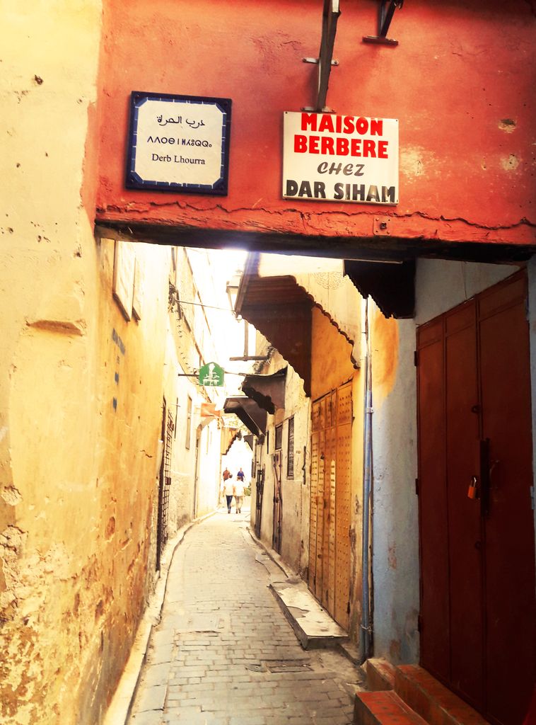 A narrow ochre alley with red doors, Arabic, Amazigh and French signs, roof projections and cables bends toward bright daylight.