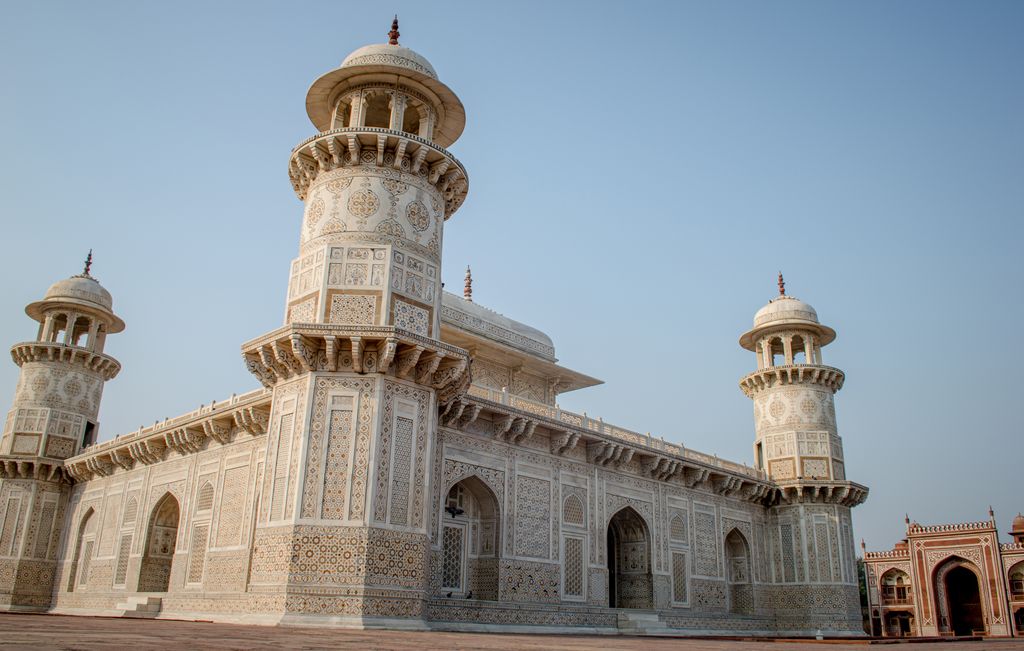 A white-marble tomb with four corner towers, deep arches and dense coloured surface patterns stands beneath a pale blue sky.