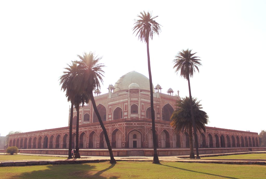 A red-and-white domed tomb rises on an arcaded platform beyond lawns, a crossing path and tall palm trees.