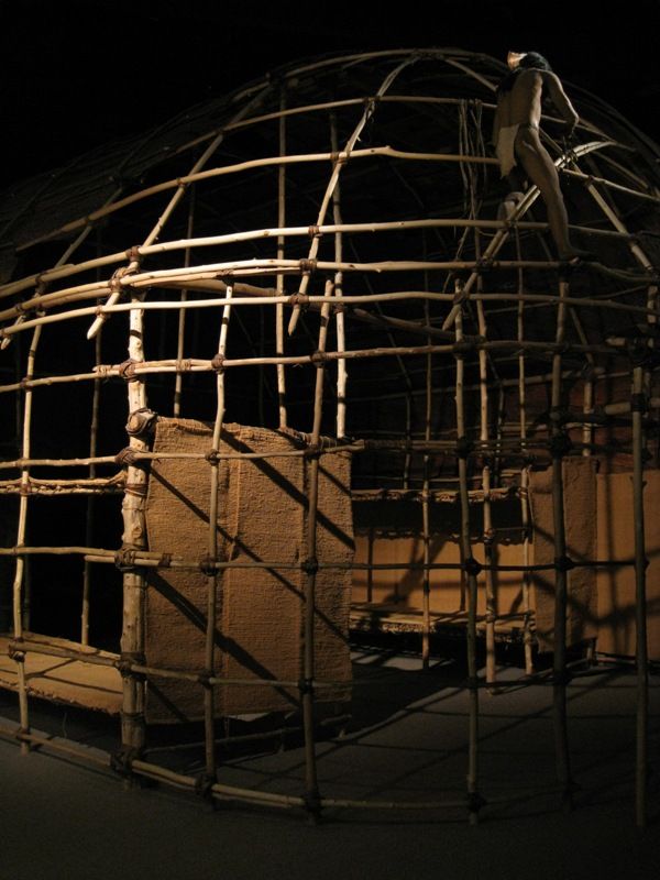 A museum display shows the curved pole frame and lashings of a longhouse replica, with partial wall panels, platforms and a mannequin inside.
