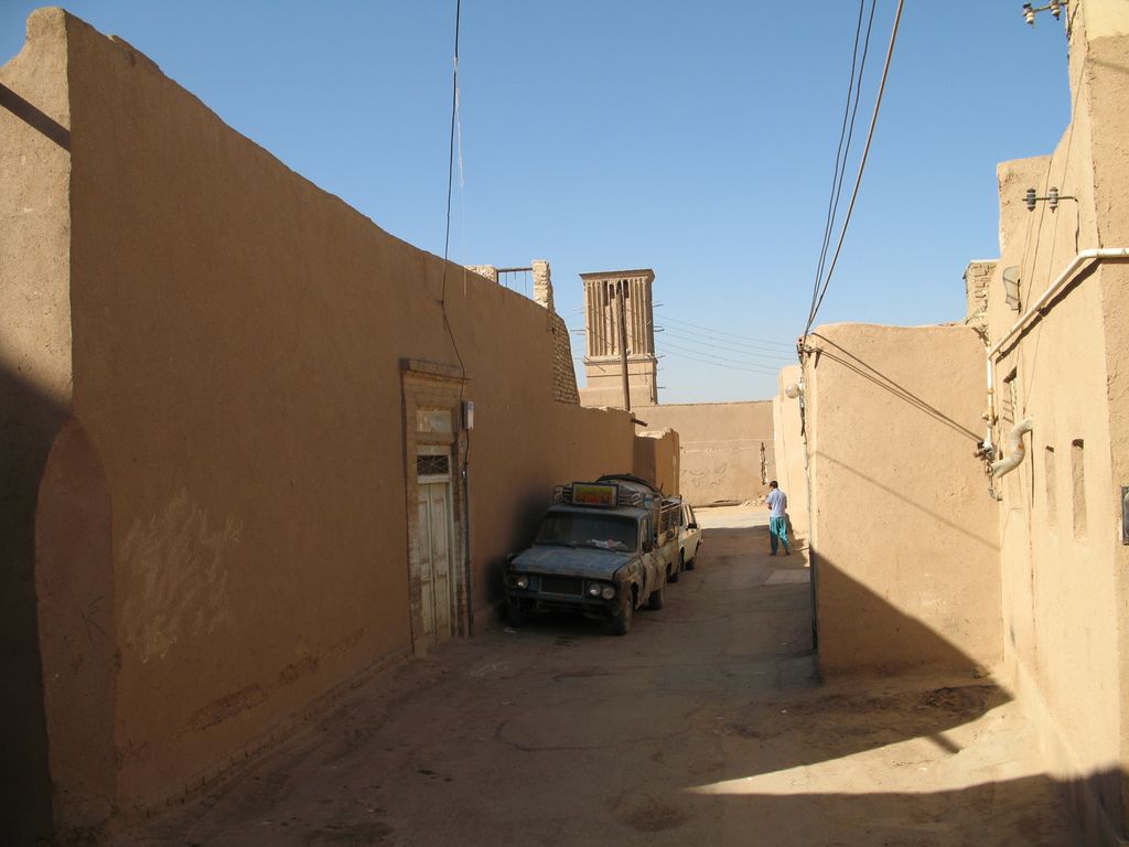 A narrow sunlit lane runs between tall ochre earthen walls toward a small windcatcher, with a parked vehicle and overhead cables.