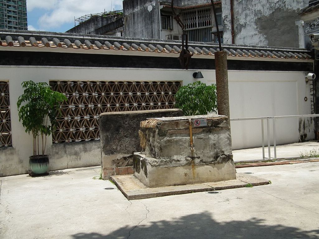 A wall with patterned curved-tile openings stands behind a stone well surround in a paved courtyard.