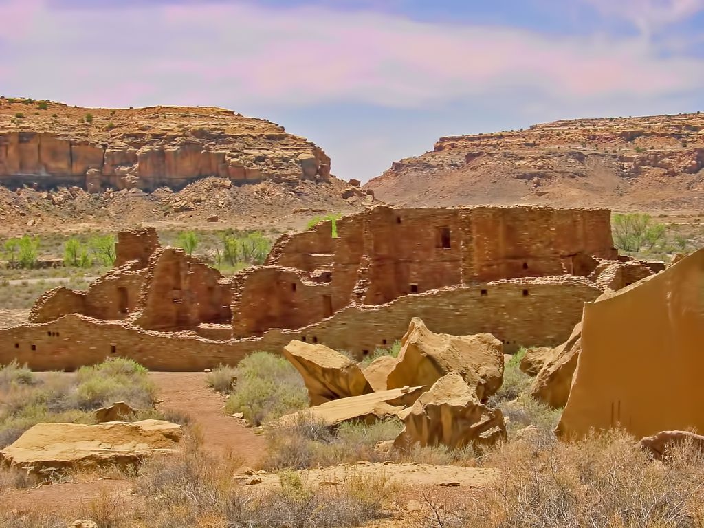 Large curving and stepped masonry ruins stand across a dry canyon floor behind angular sandstone boulders.